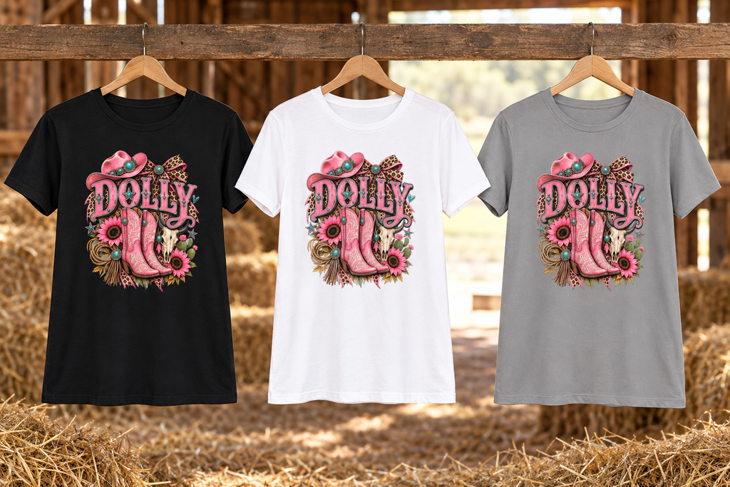 Black, white, and solid grey Dolly tees hanging loose in a rustic barn with hay